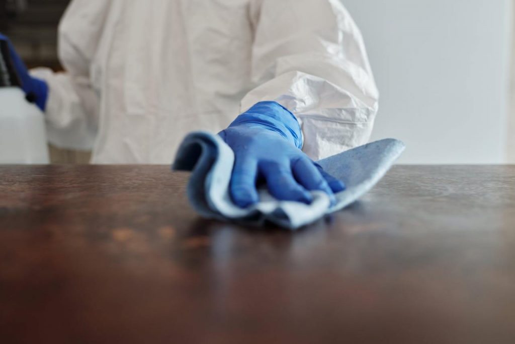 image of hands wearing a blue glove, a man is wearing a white hazmat suit and is cleaning a wooden table that's coloured brown